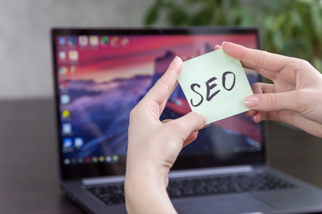 Close-up of hands peeling a sticky note that reads SEO in front of a laptop screen.